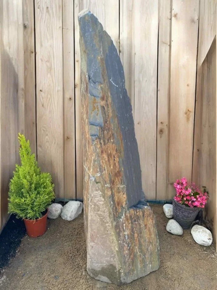 Grey slate monolith standing stone, next to stones, green bush, and pot of pink flowers against wooden fence.