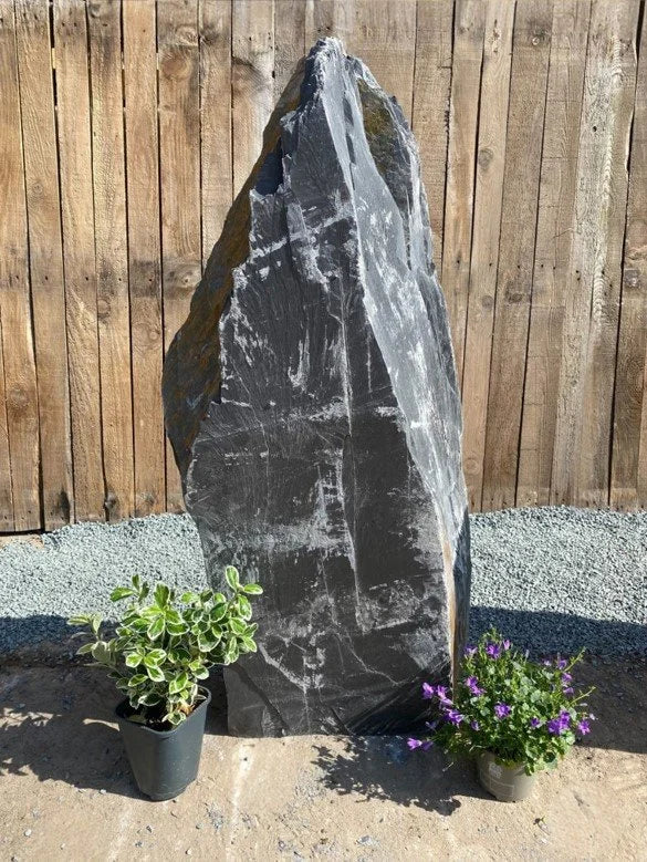 Dark slate monolith feature stone by an unknown vendor, standing tall and weathered, with small plants at the base.