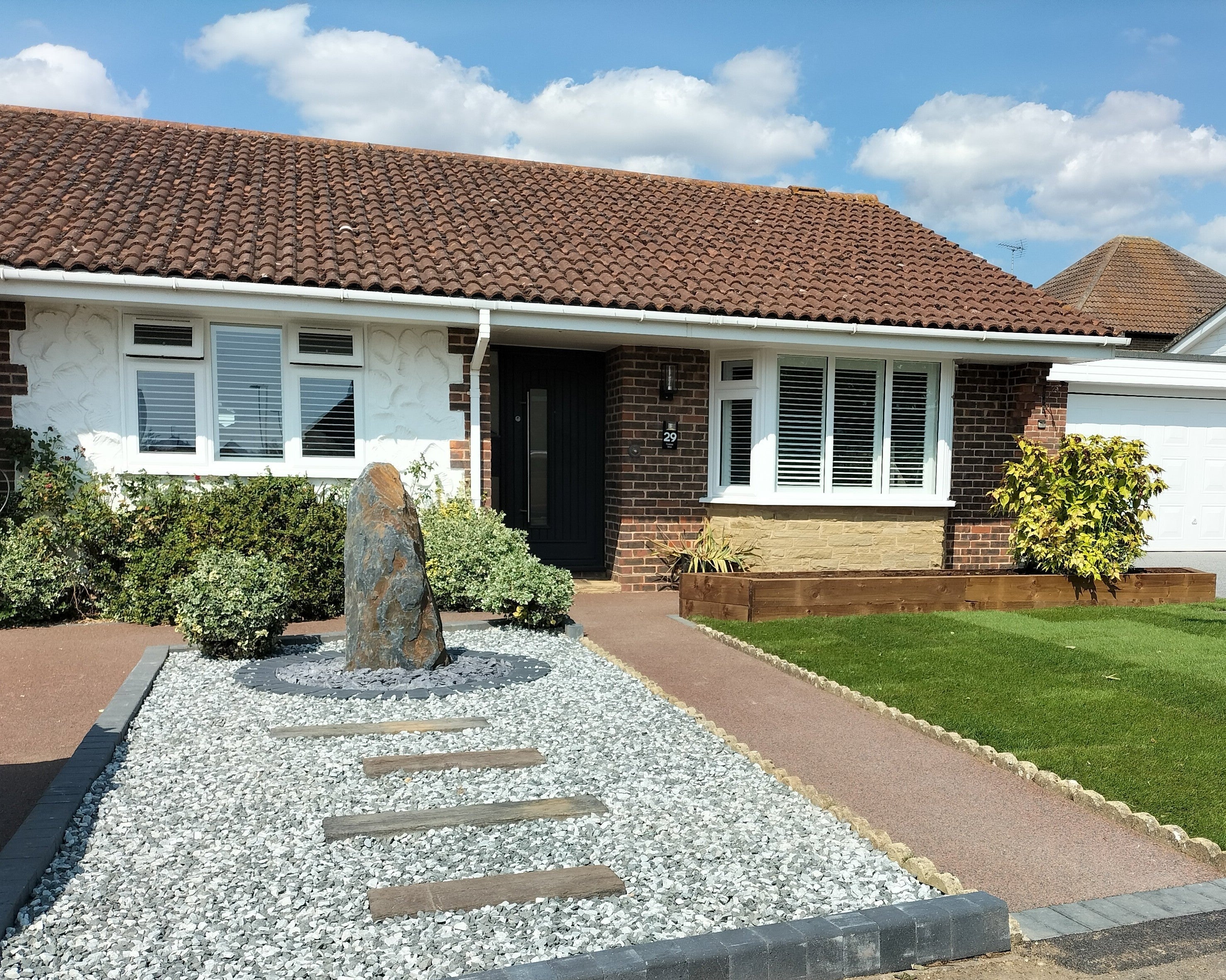 A single-story home with a red-tiled roof and brick accents, features a modern, landscaped yard with white gravel and stepping stones. Number "29" visible near the front door.