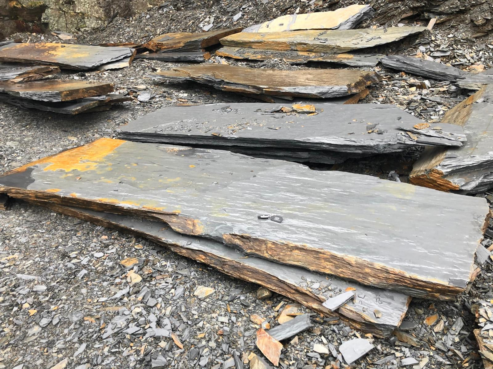 Rough grey and tan slate shards are scattered on the ground, some stacked, in a rock quarry.