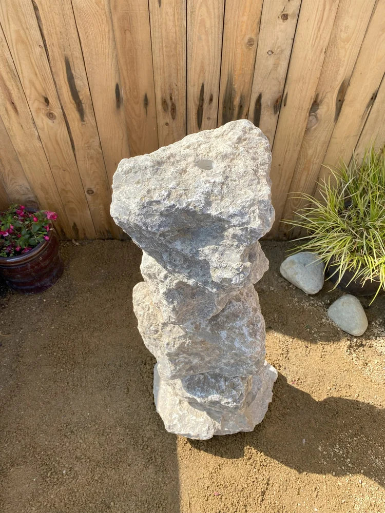 Medium grey limestone water feature, 800mm, next to plants and a wooden fence.