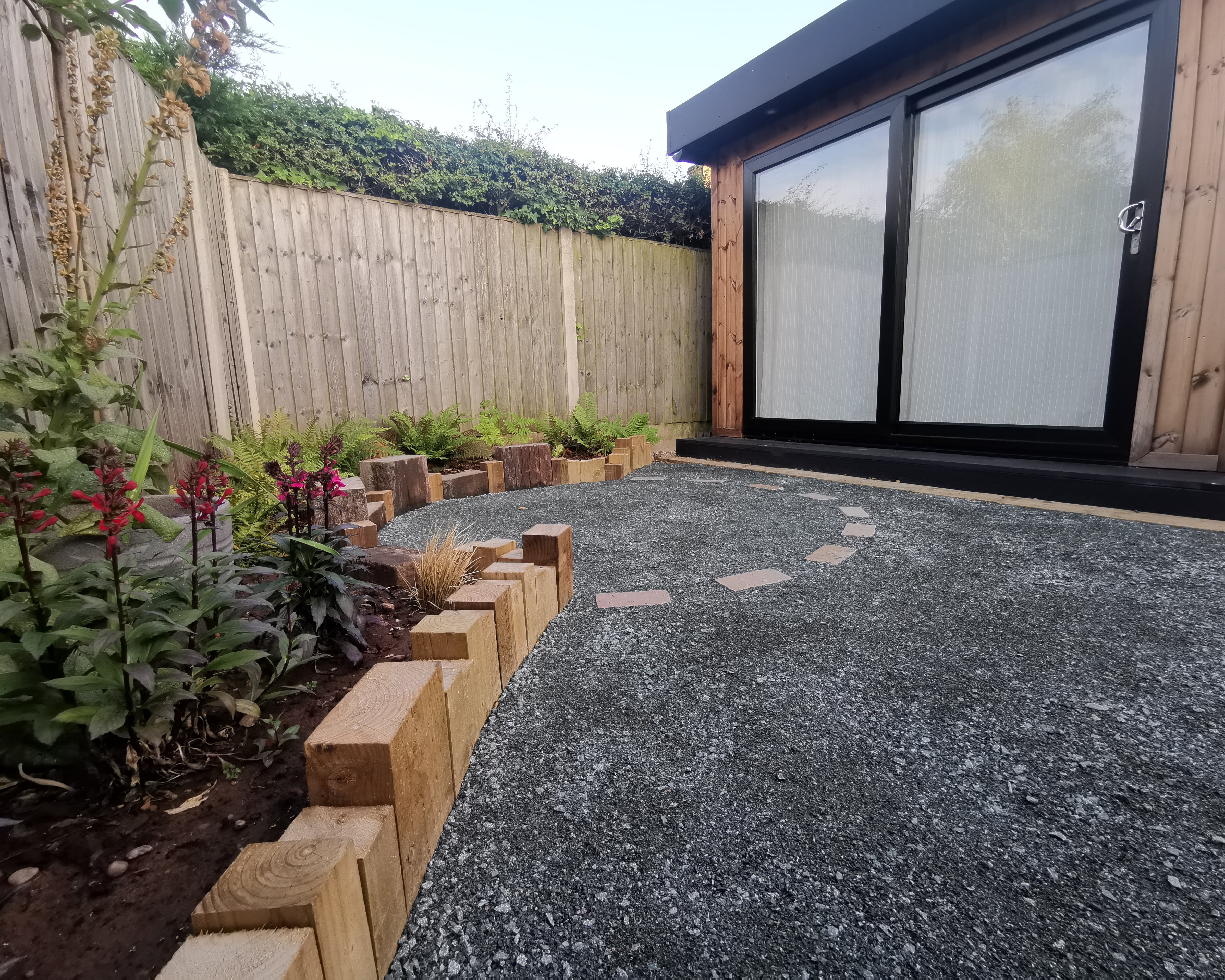 A modern garden with a wooden shed, a gravel path with stepping stones, and a flower bed with wooden edging.