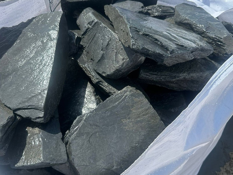 Charcoal slate decorative pond stones close-up, with layered texture, loaded in a white woven bag.