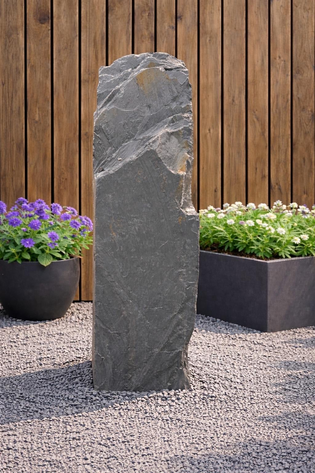 Slate monolith water feature by Water Feature 104, standing tall amongst gravel, next to potted flowers and a wood fence.