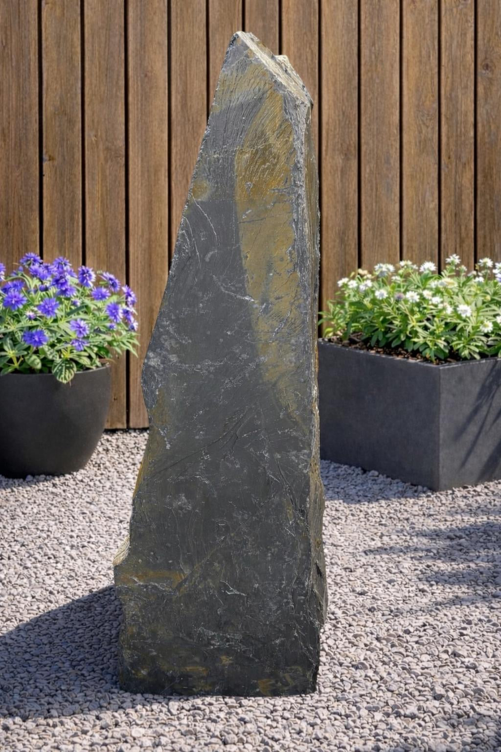 Grey slate monolith standing stone with a pointed top, standing on gravel, with planted containers in the background.