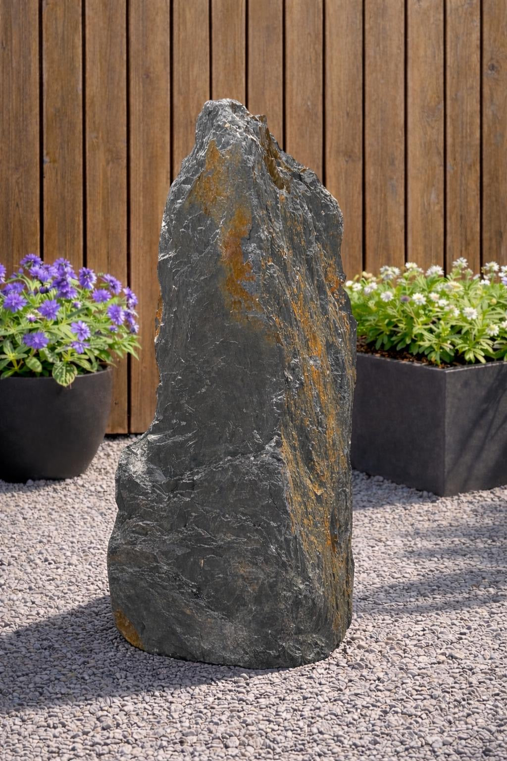Tall slate monolith water feature with orange streaks, standing on gravel in an outdoor garden setting.