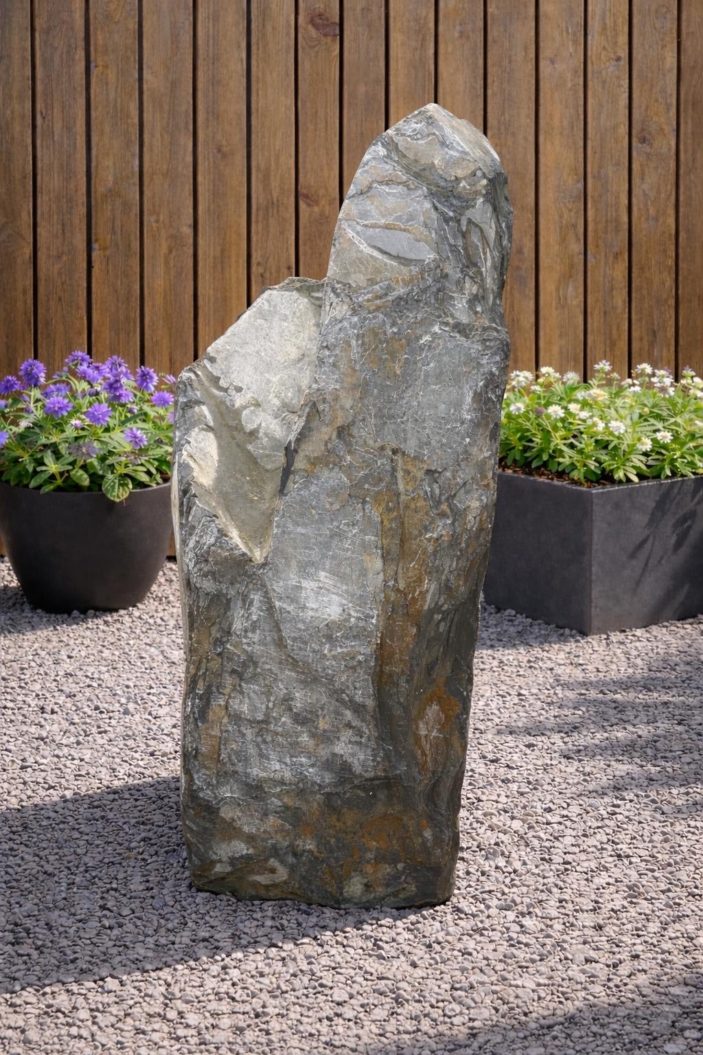 Grey slate monolith water feature from Water Feature 100, set amidst gravel and surrounded by potted flowers.