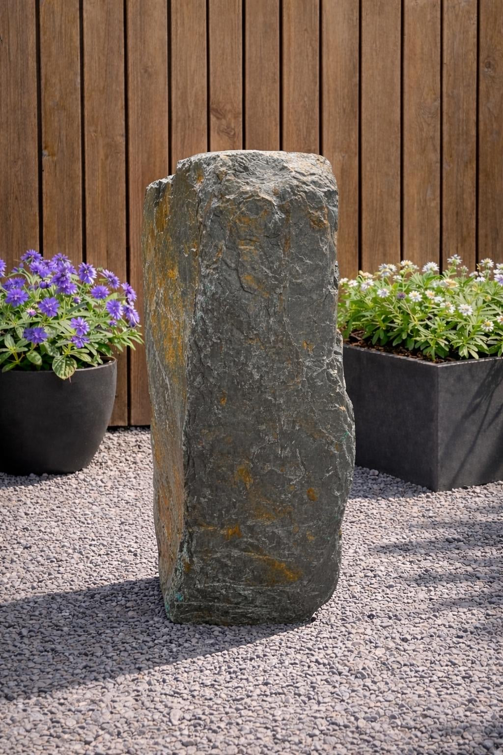 Slate monolith water feature by Water Feature 127, set amongst gravel and surrounded by potted plants.