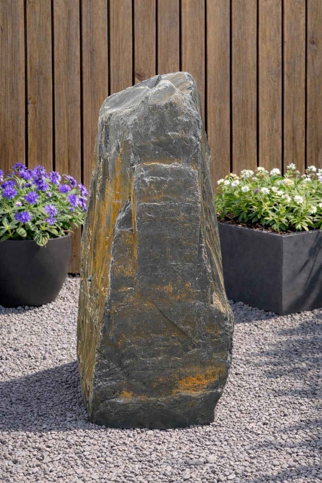 Slate monolith water feature with a rough texture, standing on gravel and surrounded by potted plants.