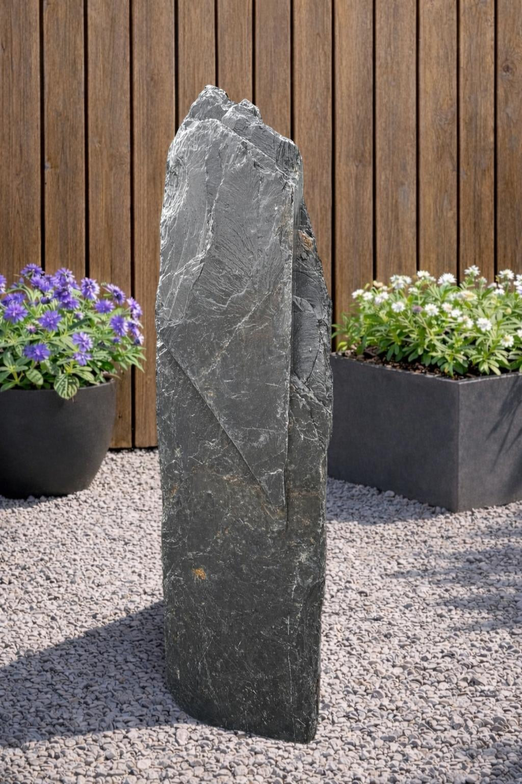 Slate monolith water feature by Water Feature 106, standing on gravel, with a rugged, layered texture.