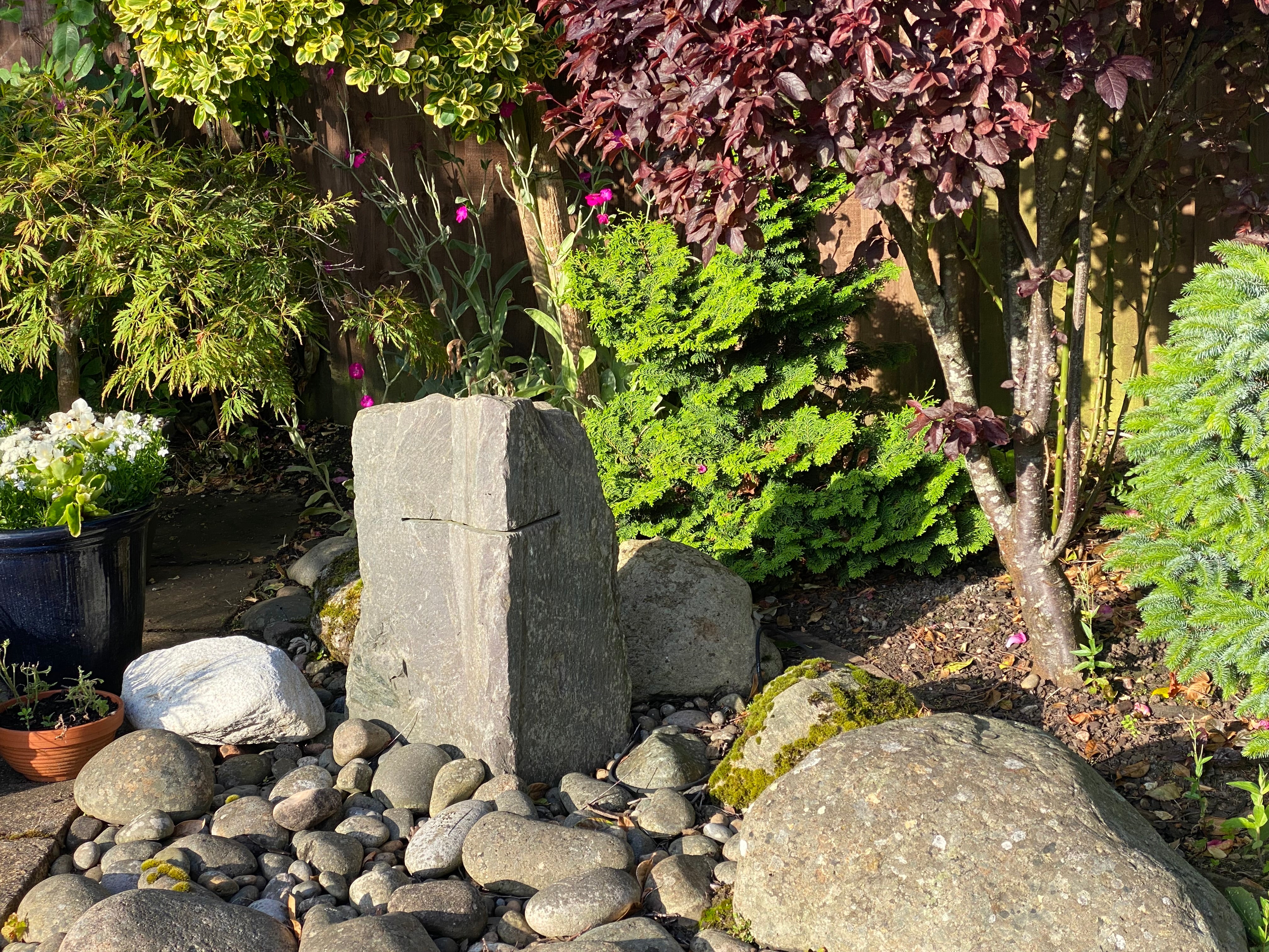 A tranquil garden scene with a stone monument in the foreground, surrounded by various plants and rocks under natural sunlight.