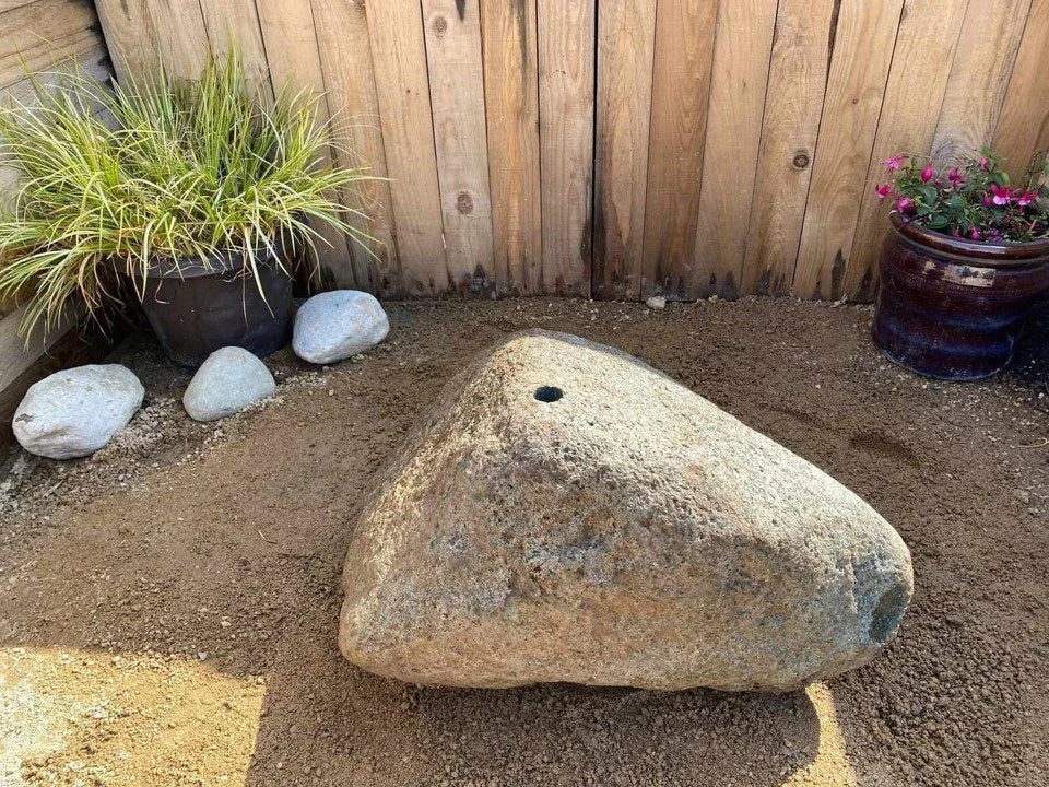 Natural drilled boulder water feature with a centered hole, placed on sandy soil near potted plants and a wooden fence.