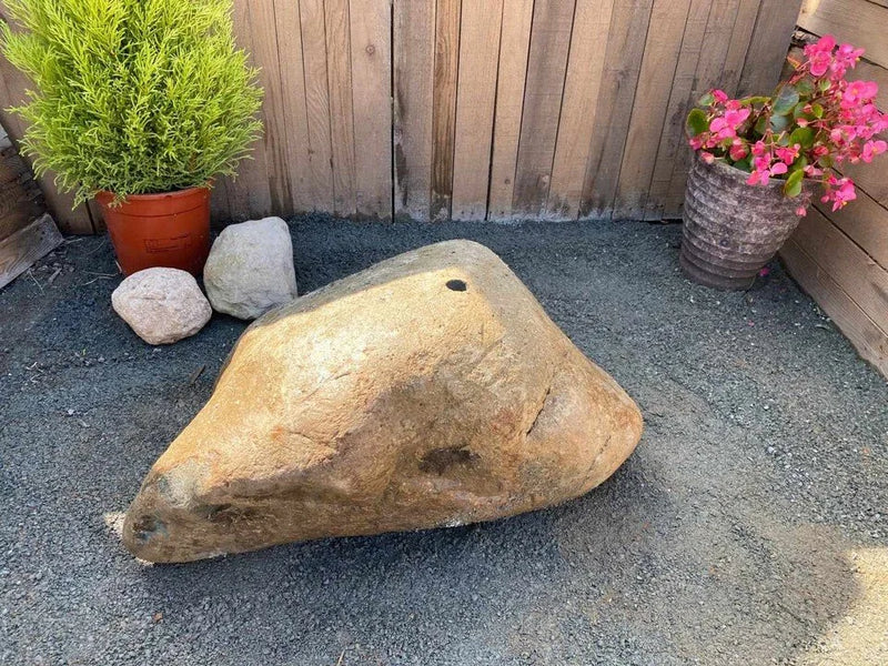 Natural drilled boulder water feature with pre-drilled hole, surrounded by gravel, next to potted plants.