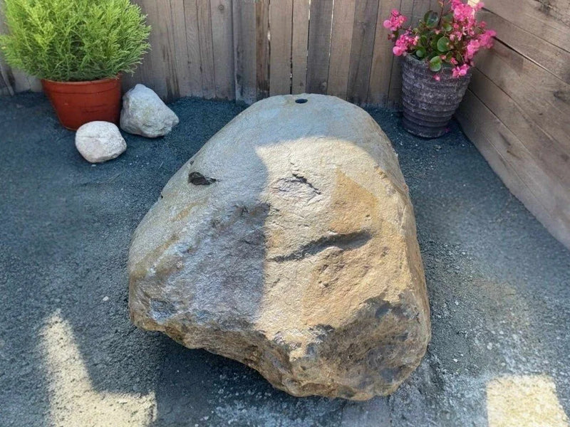Natural drilled boulder water feature, with a centered hole, sitting on gray gravel.