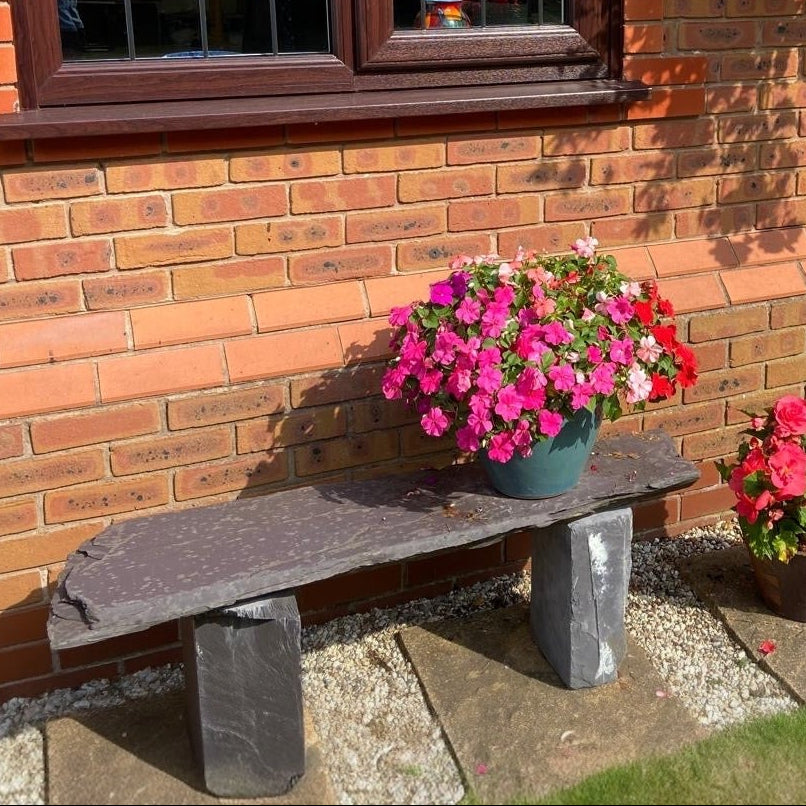A slate bench with a flower pot sits against a brick building on a sunny day. The pot is full of pink and red flowers.