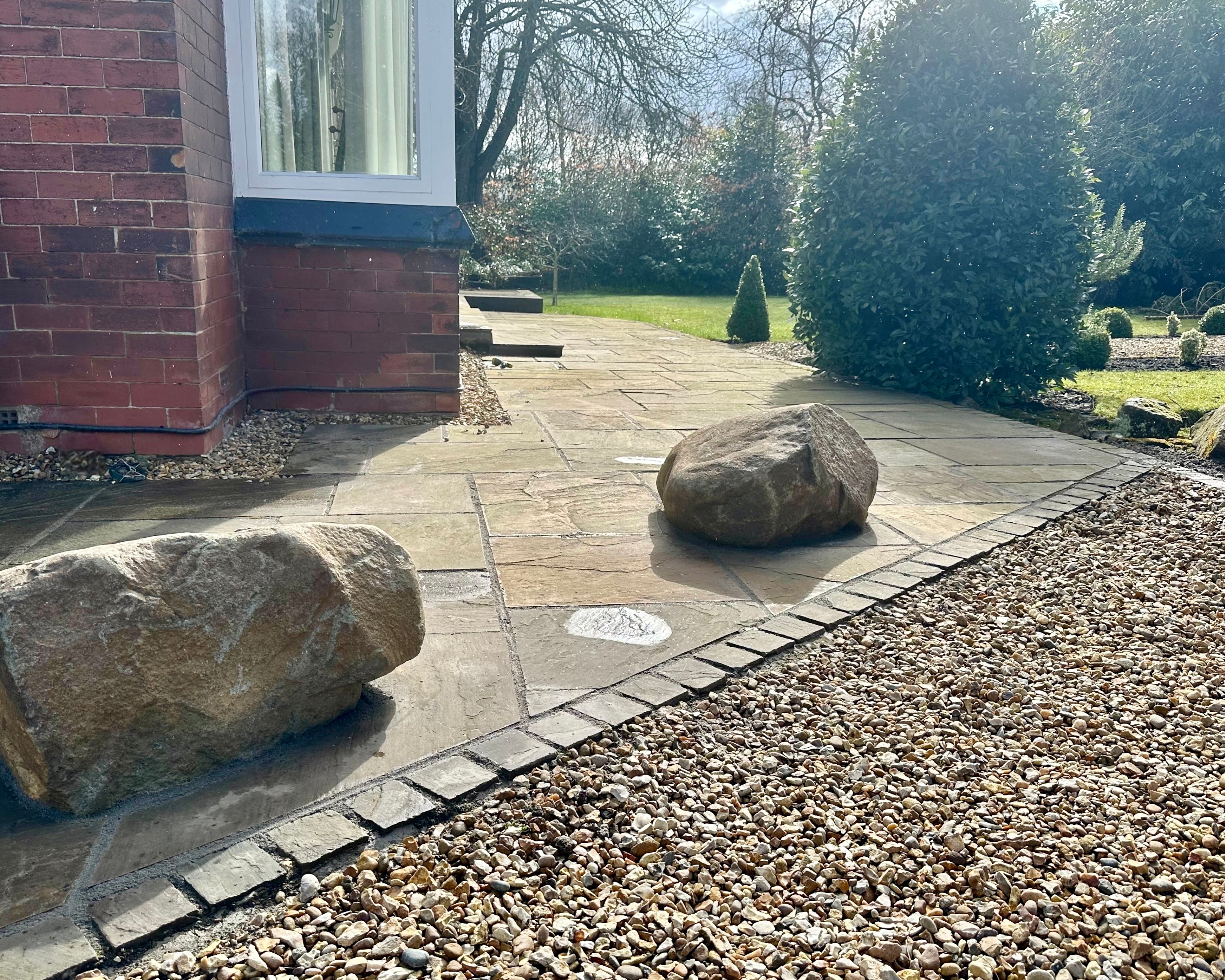 Exterior view of a paved patio with two large rocks, bordered by gravel, next to a brick building in bright sunlight.