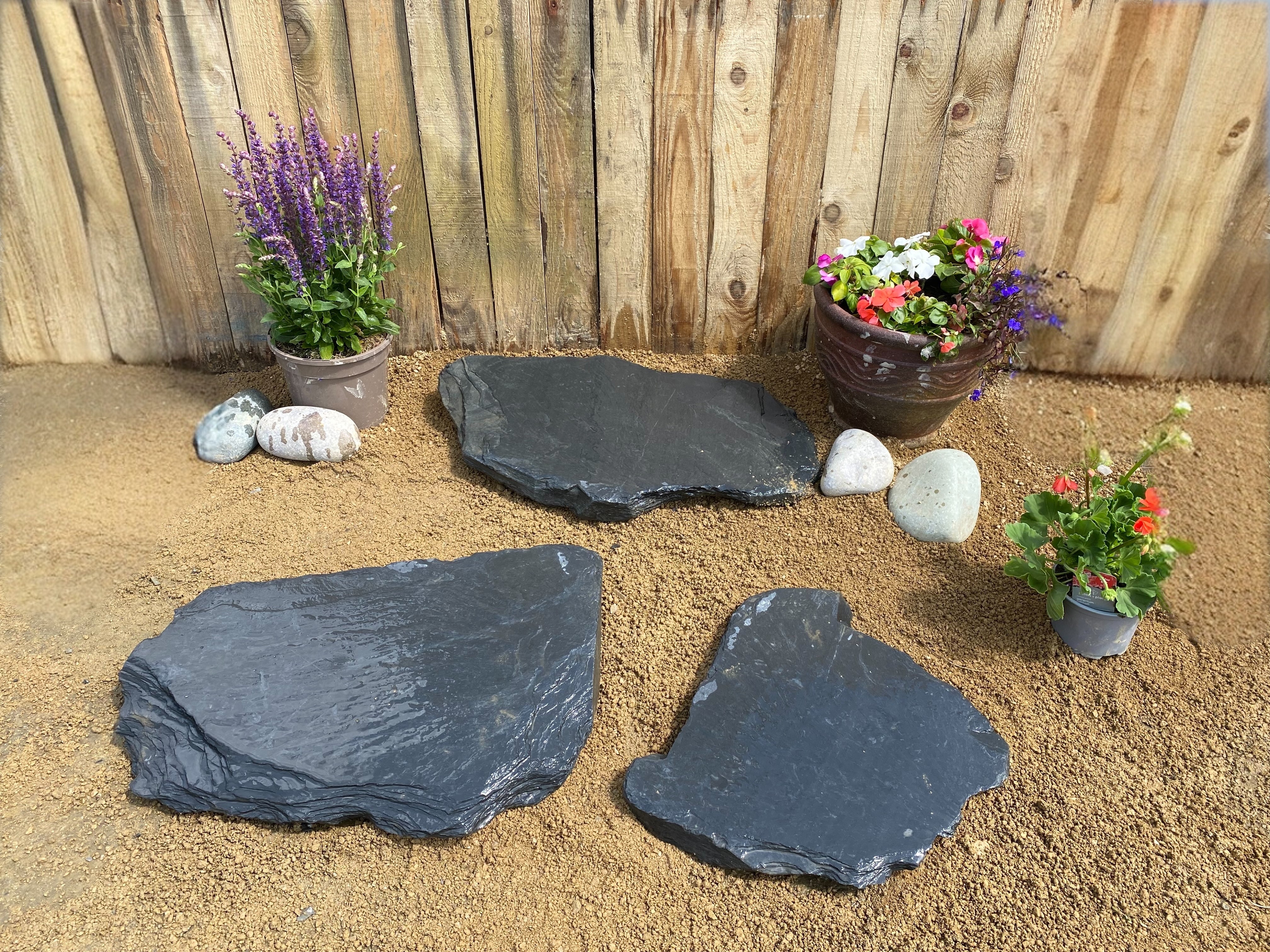 Charcoal slate stepping stones for ponds with natural irregular shapes and textured surfaces, arranged on tan gravel ground near flowering plants.