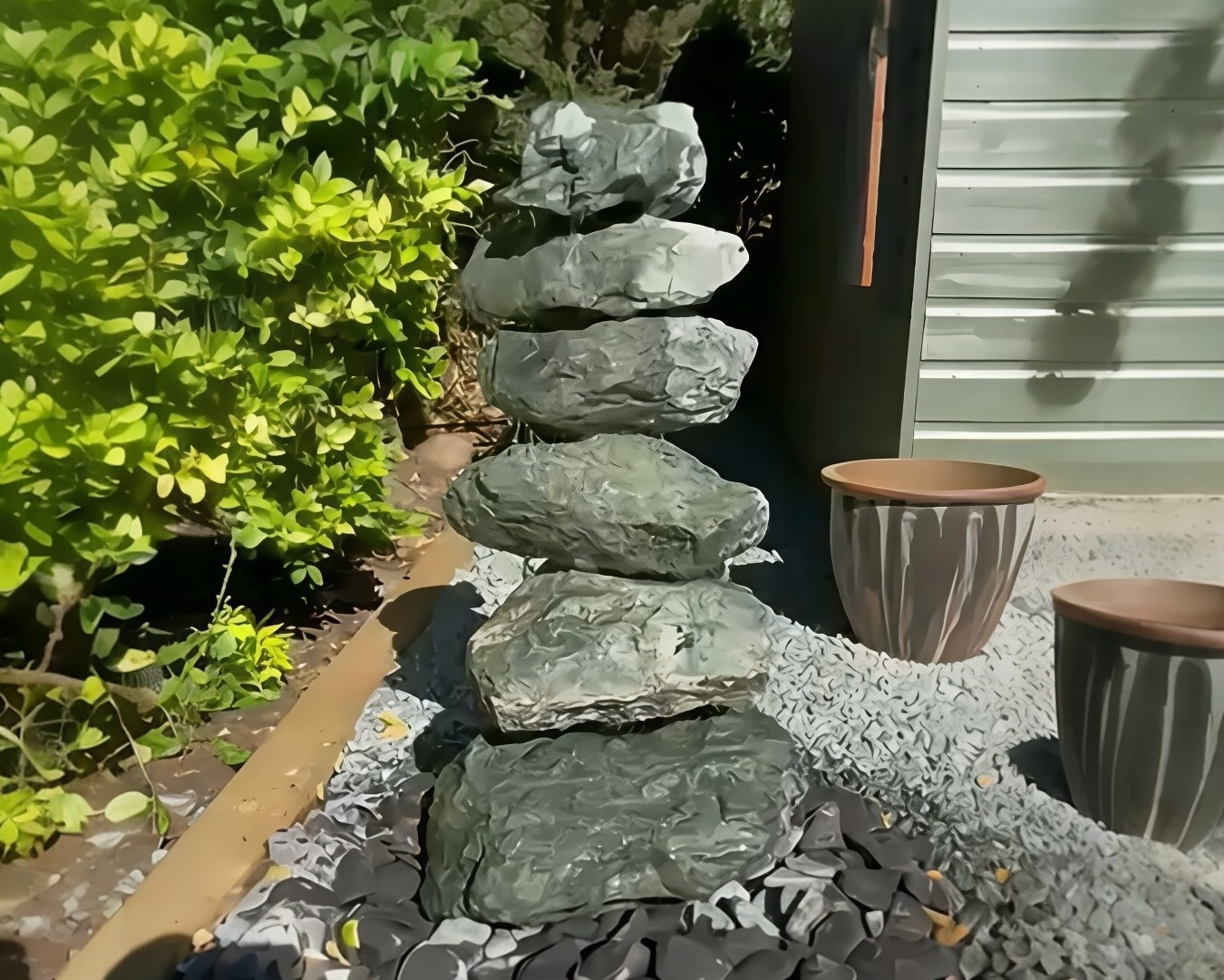 A garden scene featuring a stack of seven balanced stones, two pots, small pebbles, and green plants.