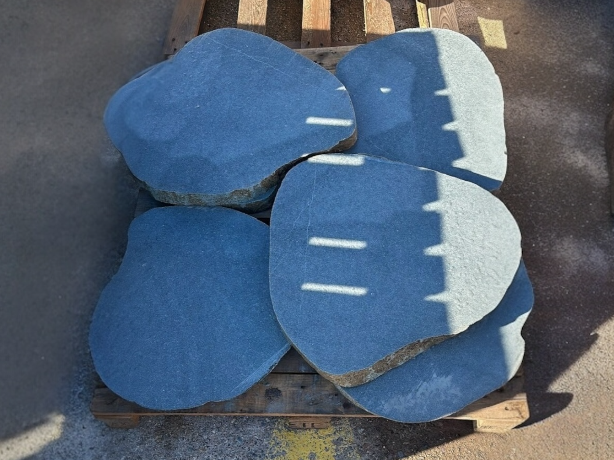Four large, irregular dark gray stepping stones stacked on a wooden pallet, bathed in sunlight casting shadows.