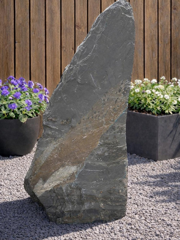 Grey slate monolith standing stone by 122 with natural textures, surrounded by pebbles and plants.