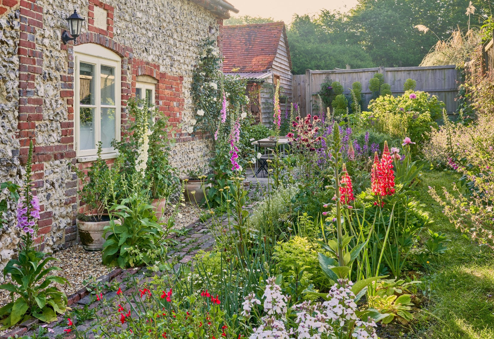 Lush cottage garden in full bloom with colorful flowers beside a stone house and garden shed. Sunlight filters through the trees.