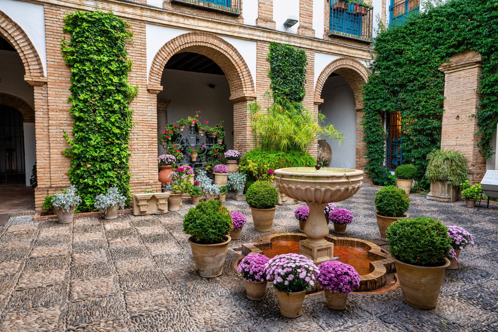 Sunlit courtyard with a stone fountain surrounded by potted plants, brick arches, and climbing greenery.