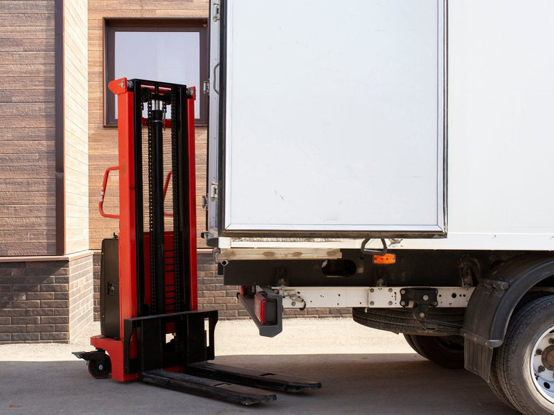 A red and black forklift sits next to the open back of a white truck in front of a building.