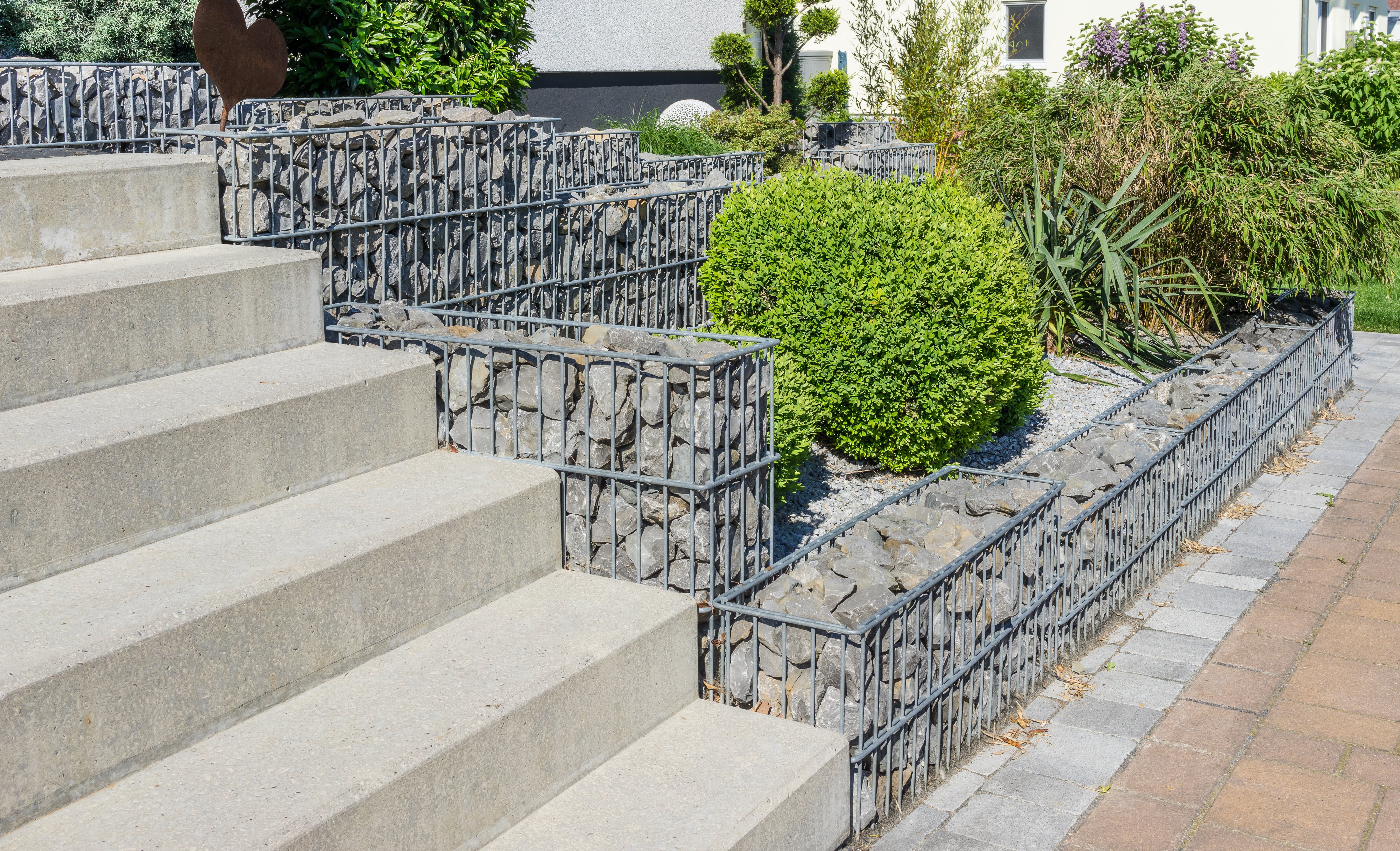 Concrete stairs ascend alongside rock-filled wire gabions, leading to greenery. The scene is bright and outdoors, highlighting the contrast of textures and colors.