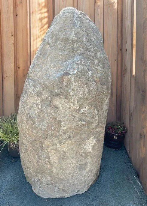 Boulder monolith standing stone by G3, tall and roughly textured, stands on a gray surface next to a wooden fence.