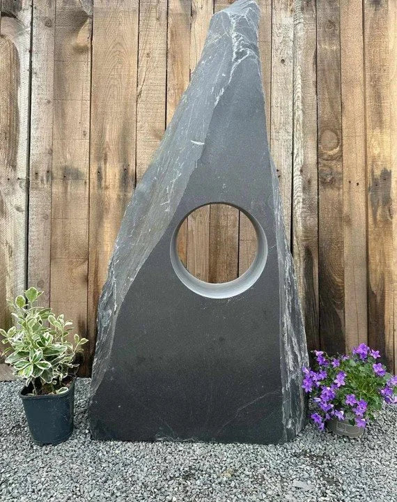 Grey slate hollolith water feature (900mm) with a circular hole, placed on a pebbled surface, with wooden fence background.
