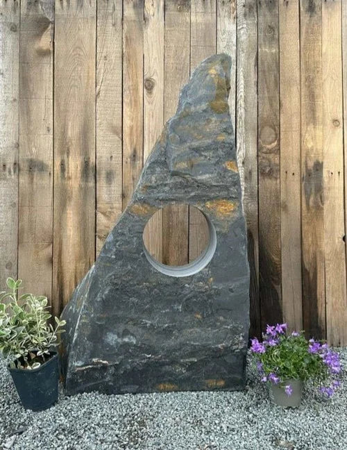 Grey slate hollolith water feature with a central round hole, by Ambient, next to potted plants.