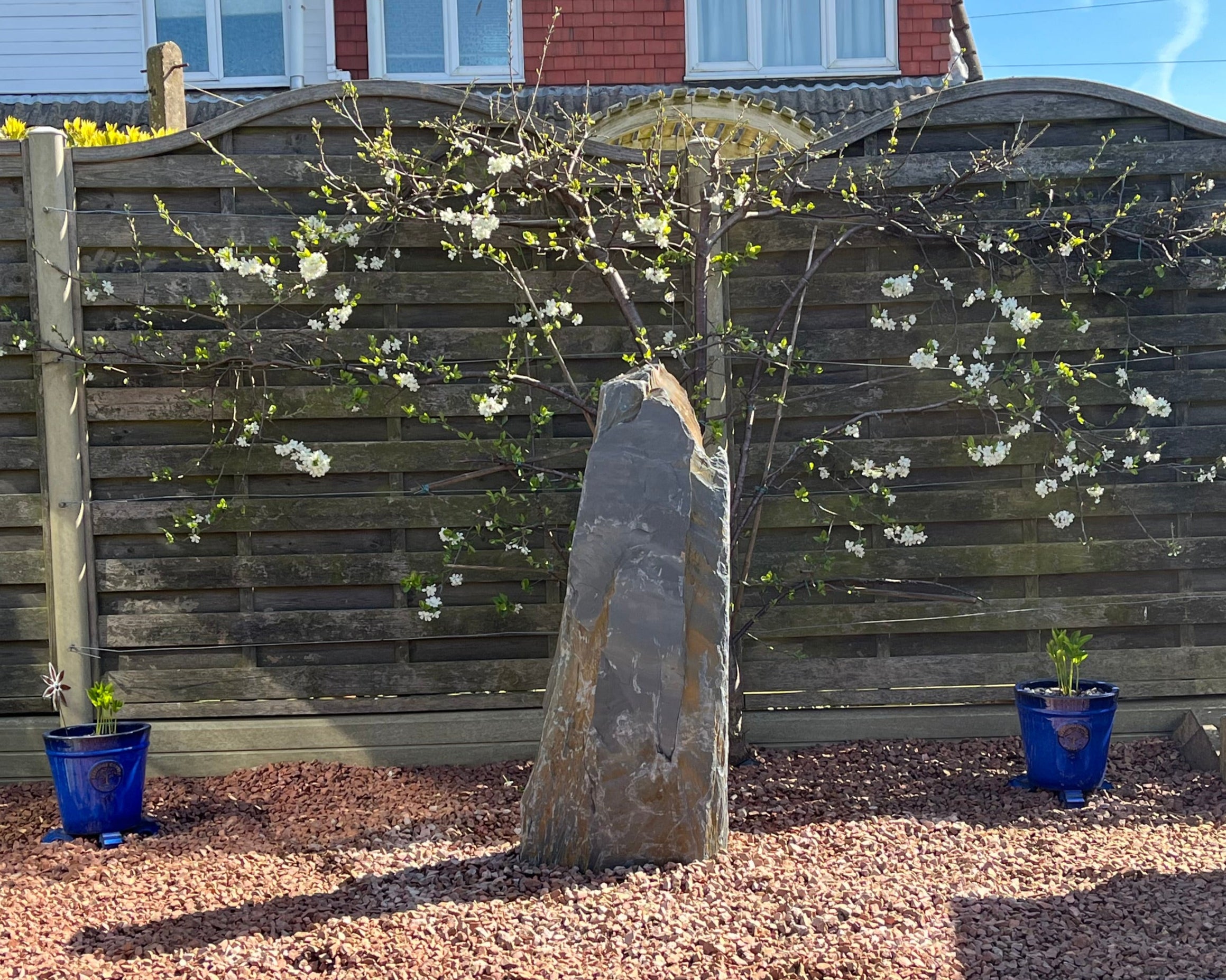 Outdoor garden scene: a tall stone sculpture stands among red gravel, framed by a blossoming tree and a weathered wooden fence. Two blue planters flank the scene.