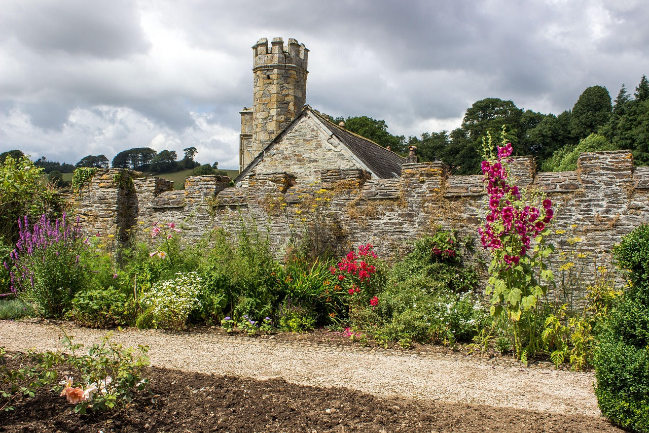 A walled English garden with a stone tower rising in the background on an overcast day. Various flowers line the gravel path.