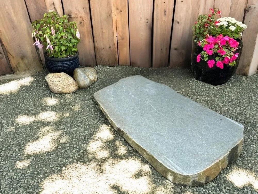 Sawn large stepping stone for modern gardens with a grey stone texture, among decorative gravel and plants.