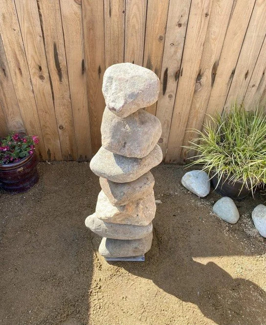 Medium stacked stone pebble tower from an unbranded vendor, set outdoors with plants.