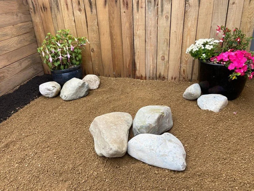Medium natural pond boulders, 200-350mm, scattered across sandy soil near potted plants and a wooden fence.