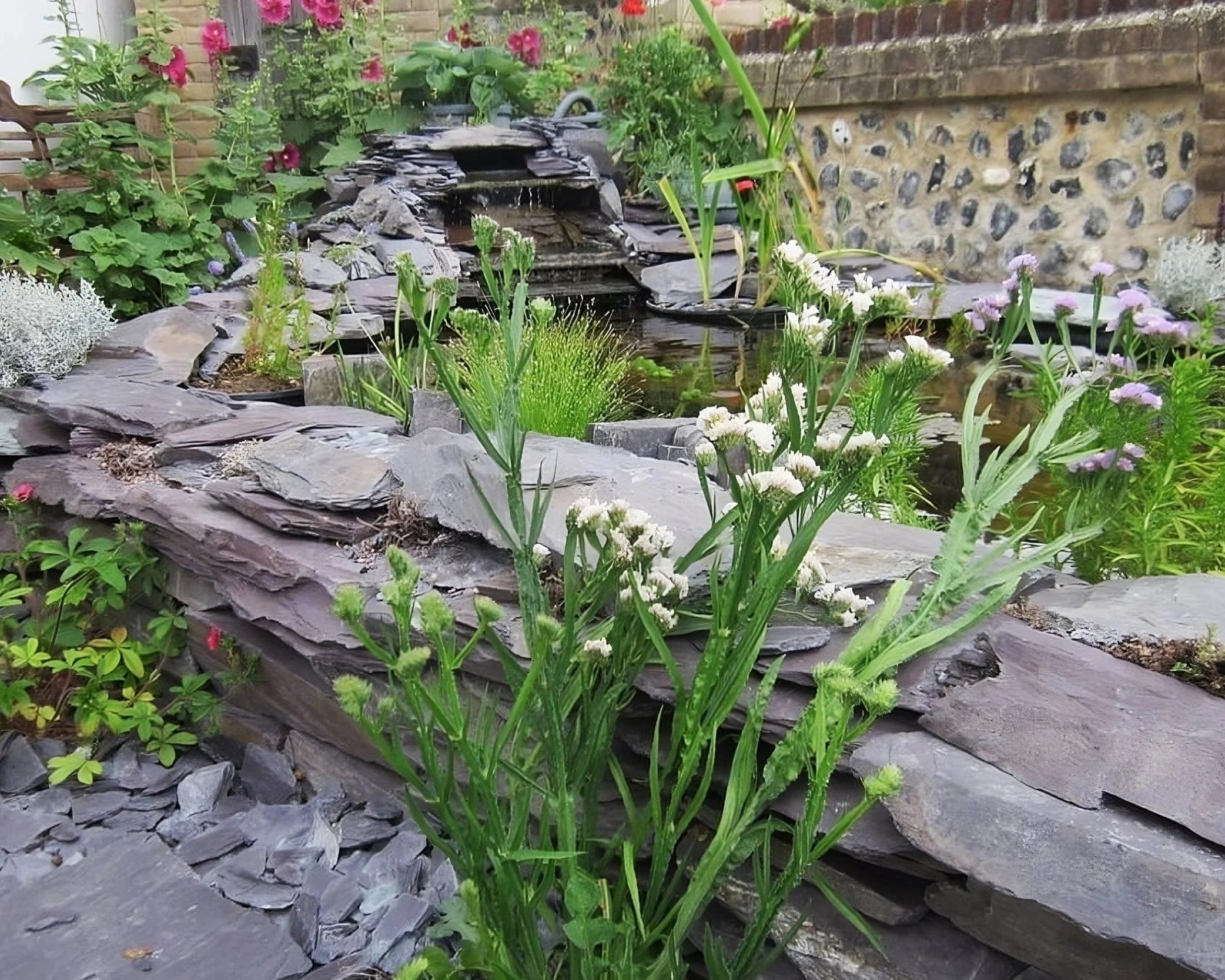 A vibrant garden scene with a stone waterfall cascading into a pond, surrounded by various green plants and colorful flowers.