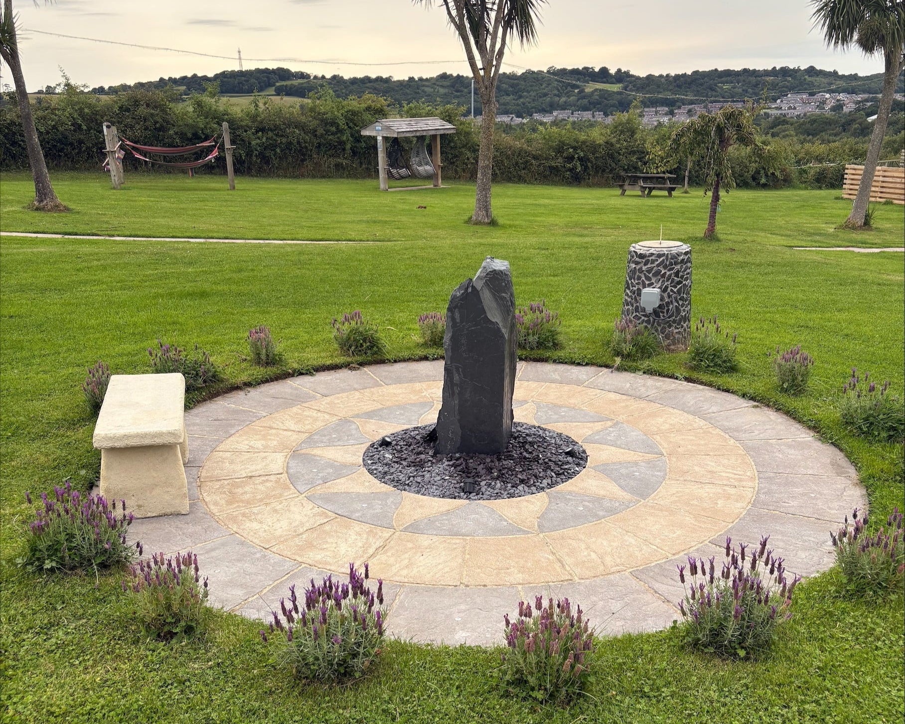 A circular stone patio with a tall, dark rock fountain in the center sits on green grass, surrounded by lavender bushes. A bench is to the left.