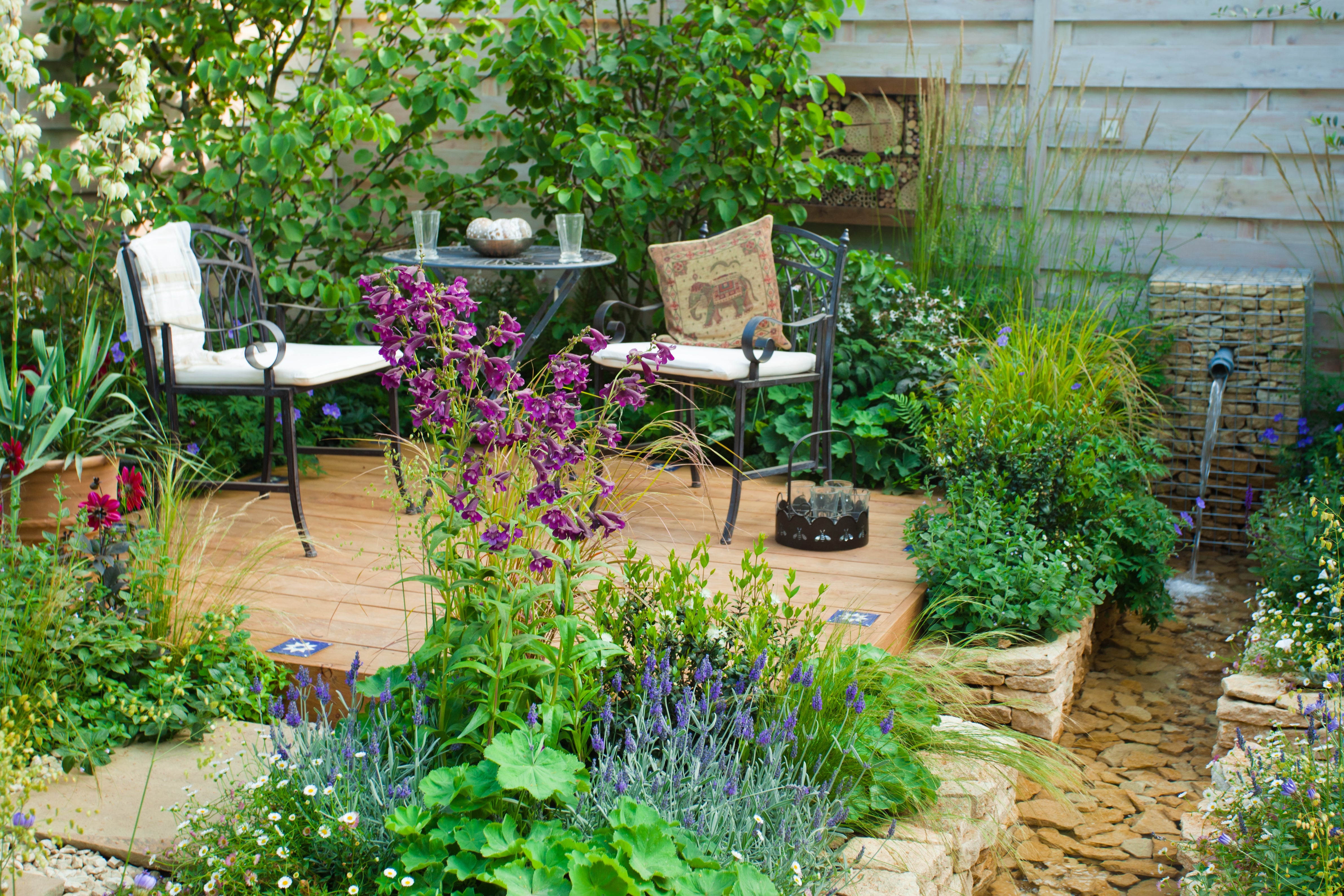 Lush garden with a wooden deck featuring two chairs, a table with glasses, and a stone water feature. Purple flowers add color to the scene.