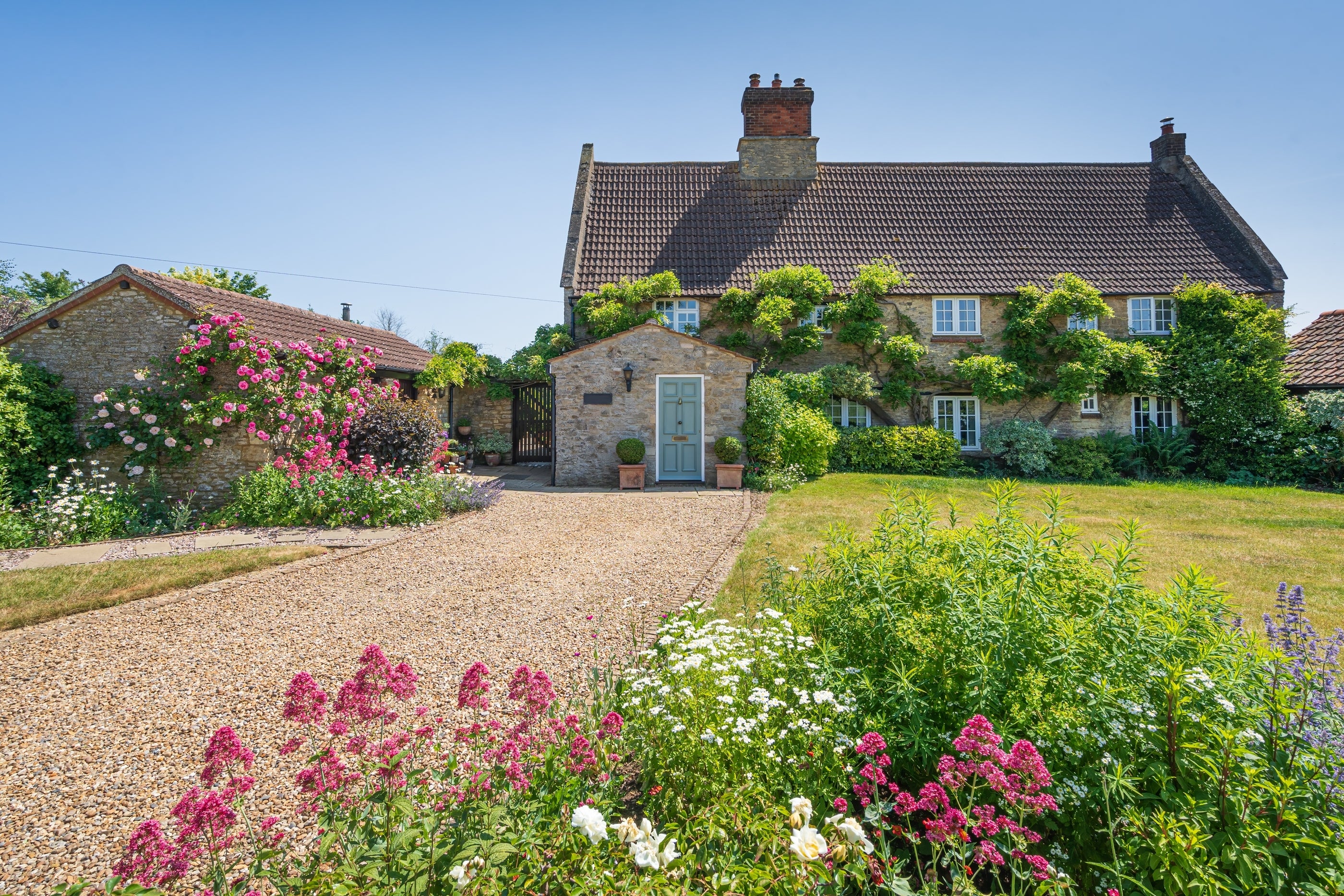A picturesque stone house with blooming garden on a sunny day. Gravel driveway leads to the front door. Lush greenery surrounds the property.
