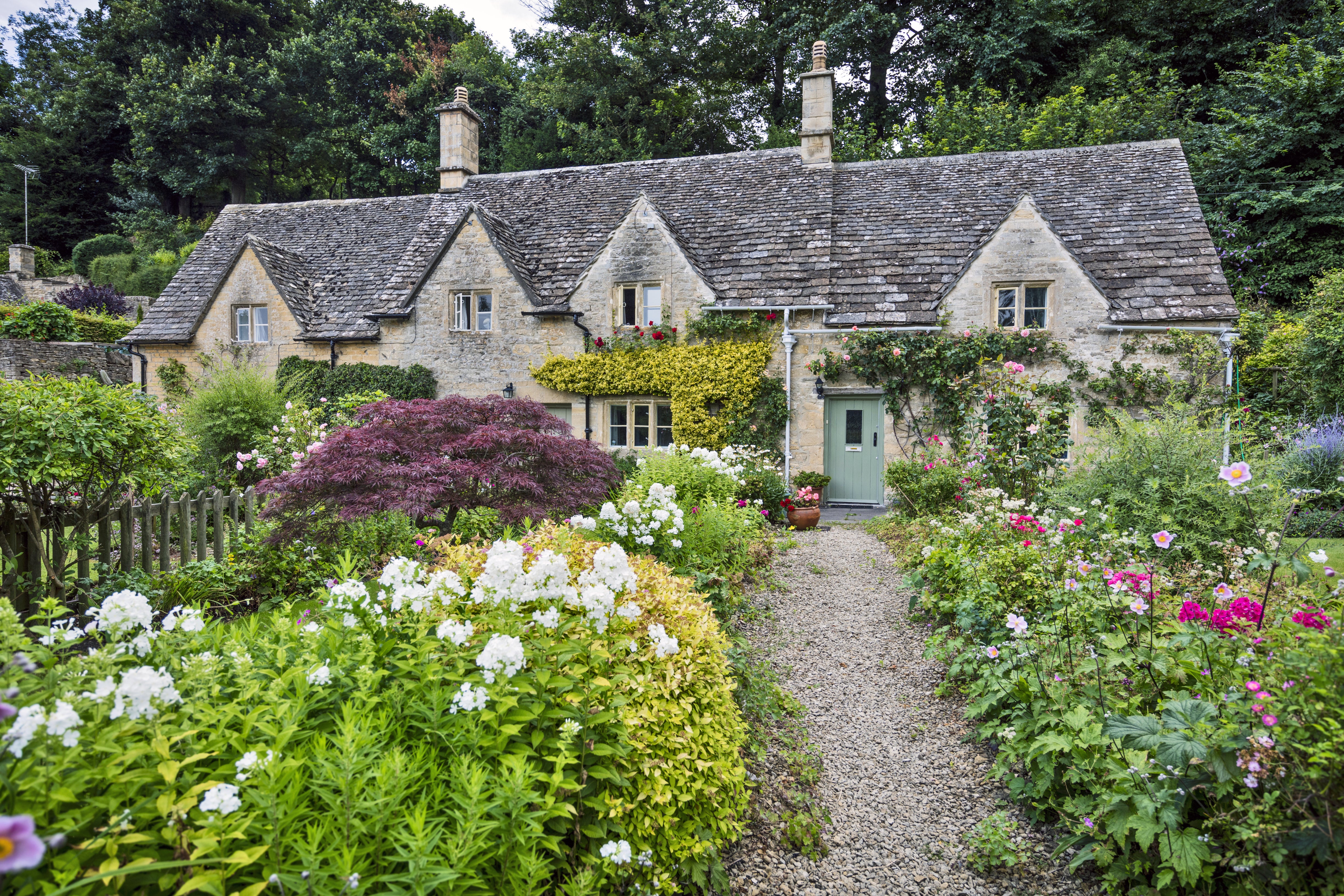 A stone cottage with a green door sits amid a vibrant garden with white and pink flowers. A gravel path leads to the entrance.