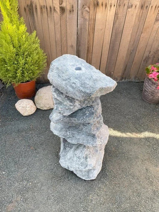 Small green granite water feature (600mm) with layered stones, set in an outdoor gravel garden with plants.