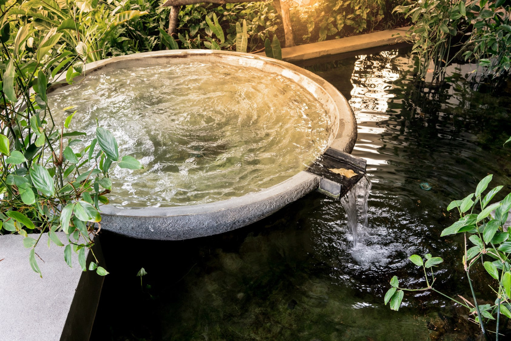 Overhead shot of a serene stone fountain, overflowing into a dark pond surrounded by lush greenery. Sunlight creates a tranquil atmosphere.