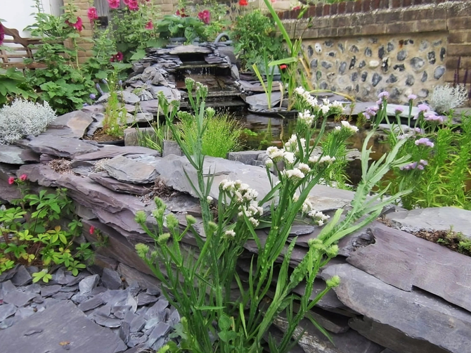 A tranquil slate-lined pond with a small waterfall, surrounded by lush greenery and blooming flowers, creating a serene garden oasis.

