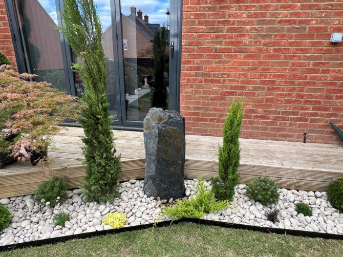 A calming garden water feature made of a tall, dark stone is surrounded by lush plants and smooth white stones.