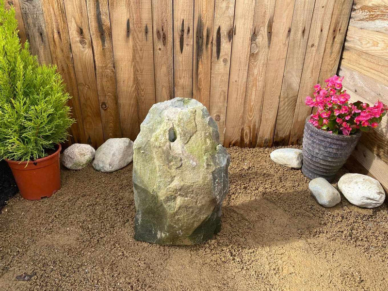 Gray slate monolith water feature from GSM, with a hole detail, surrounded by pebbles and plants.