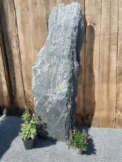 Grey slate monolith standing stone by Standing Stone, with a textured surface, standing on gravel near potted plants.