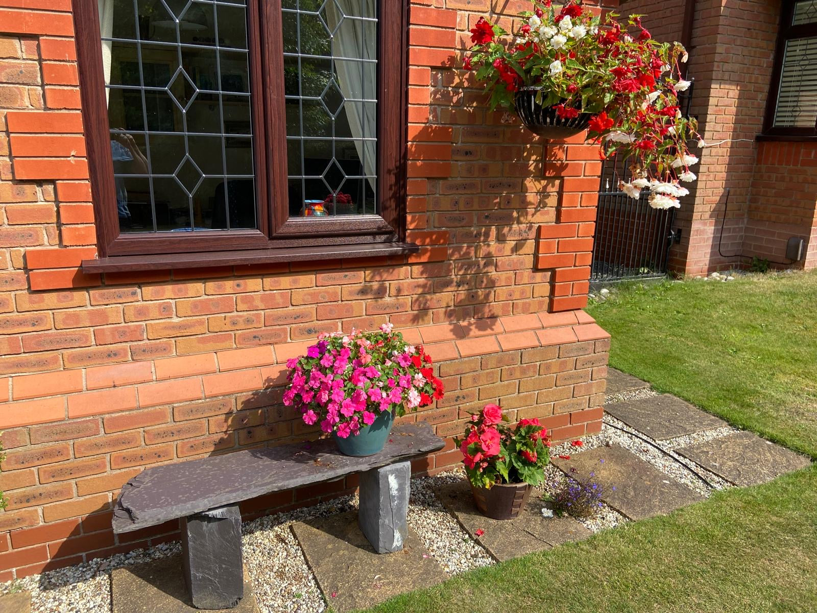 A sunny outdoor scene of a brick building with a window and hanging flower baskets. A stone bench with potted flowers sits in the foreground.