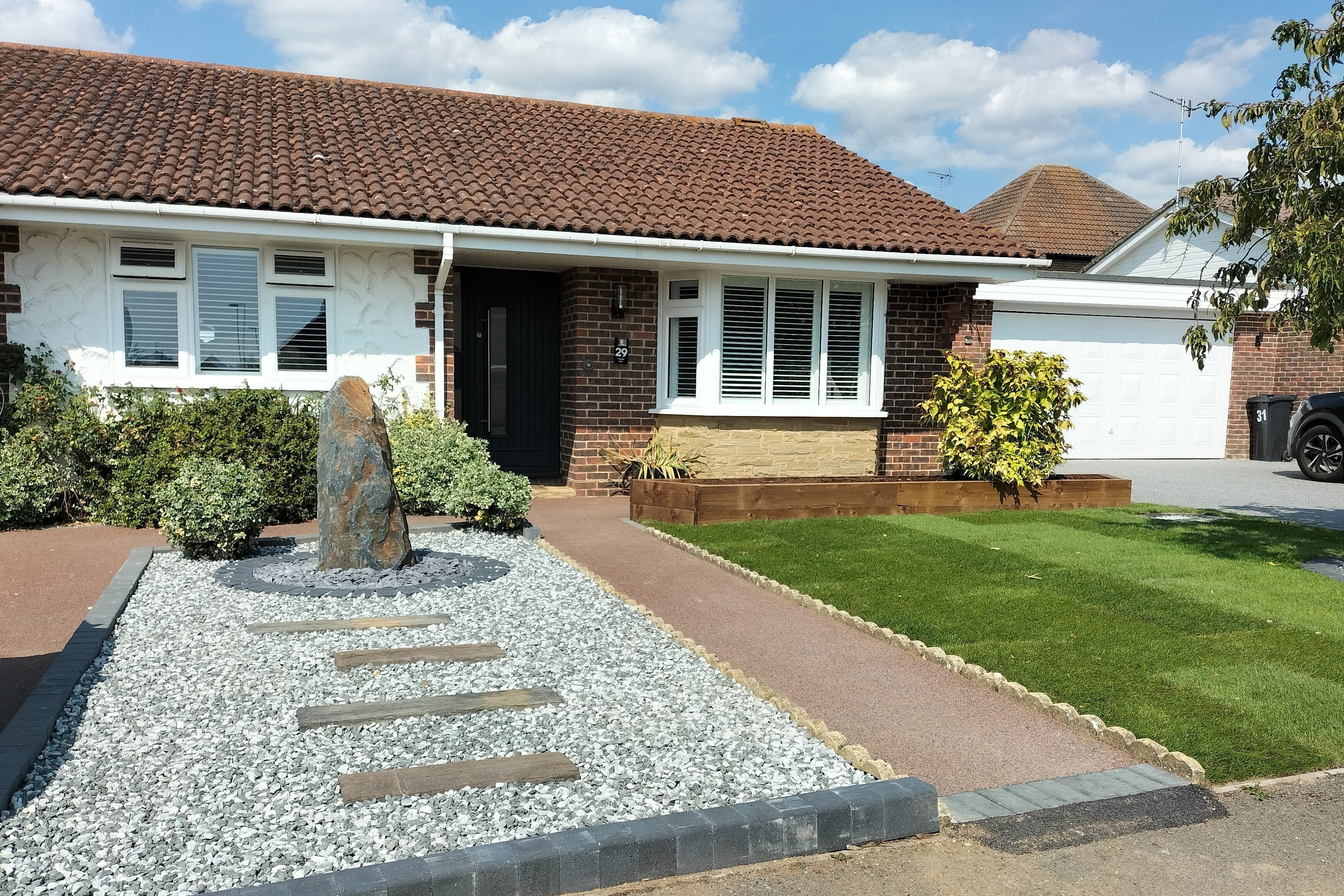 A well-maintained front yard with a bungalow; stone sculpture, gravel, and stepping stones are visible. A pathway leads to the front door.