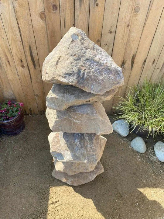 Large golden granite water feature with stacked rock design, near potted plants and a wooden fence.
